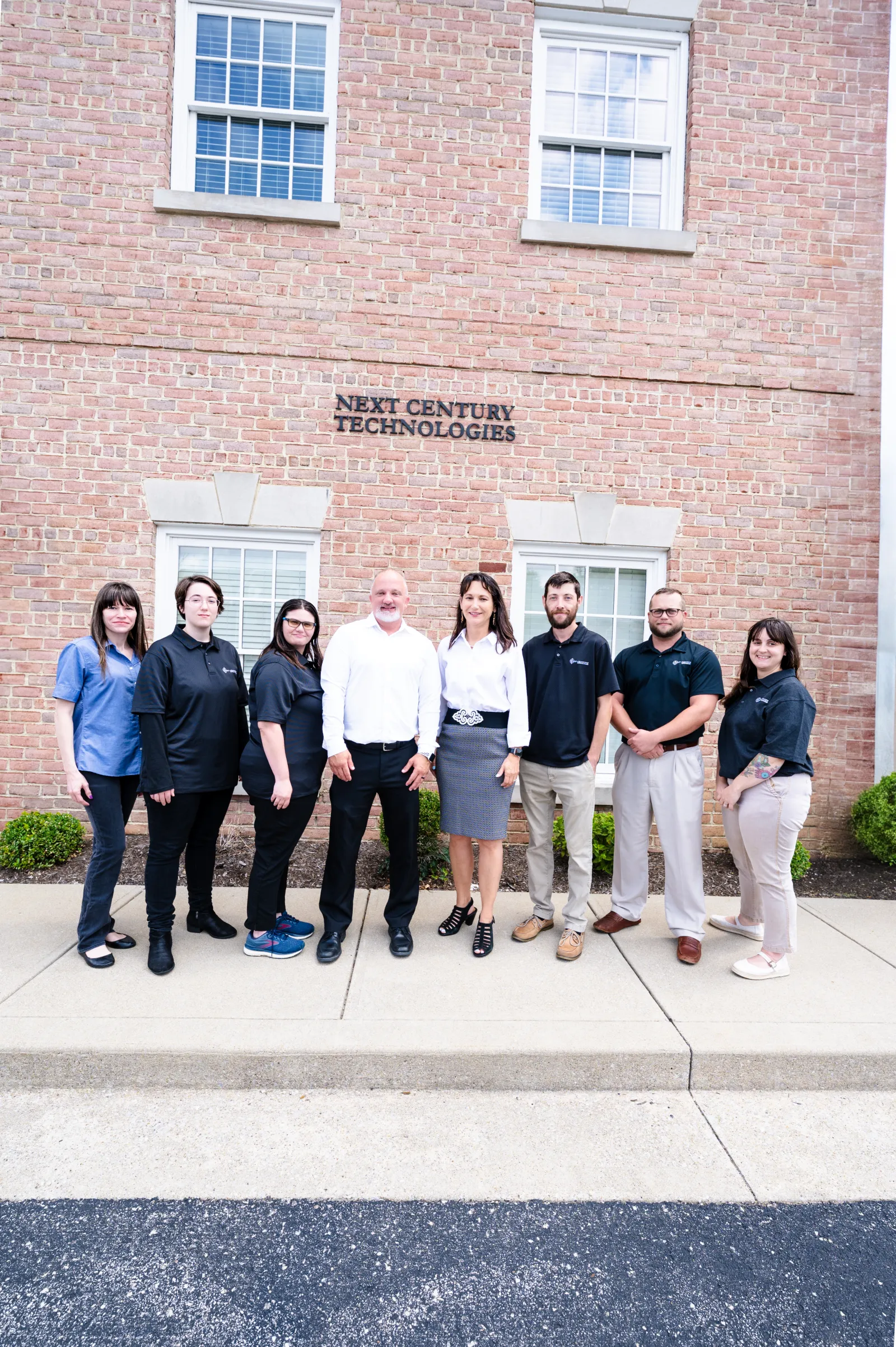 Group of eight professionals standing outside Next Century Technologies office building with brick facade.